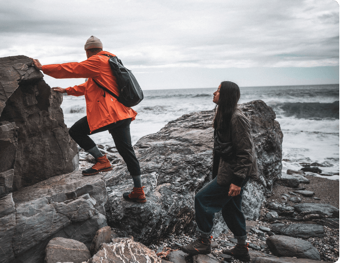 Two people climb seaside rocks in outdoor gear as waves crash under a cloudy sky.