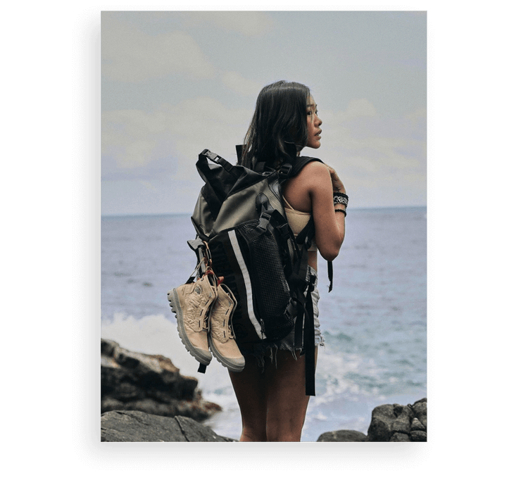 Person with long hair and backpack stands on rocky shore, gazing at sea under a cloudy sky.