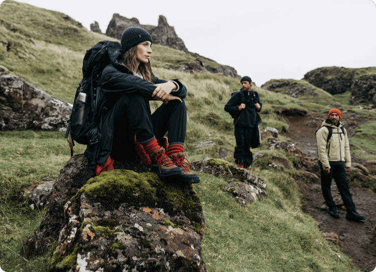 Three hikers on rocky hillside; one sits on mossy rock, two walk in the background under overcast sky.