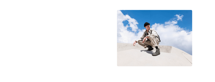 Person in beige outfit, black beanie, and boots squats on concrete structure under blue sky with clouds.