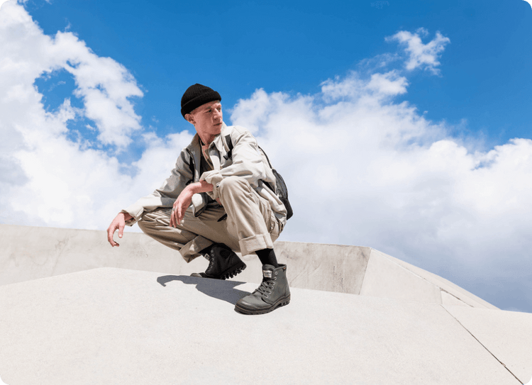 Person in beige outfit, black beanie, and boots squats on concrete structure under blue sky with clouds.