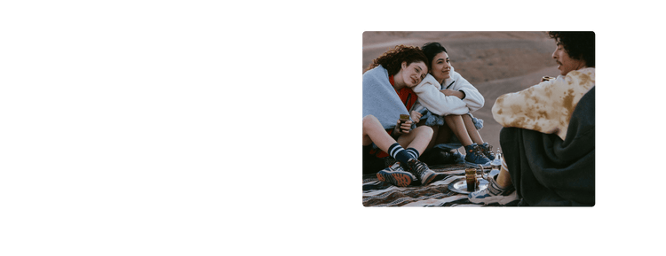 Three friends sit on patterned blanket outdoors, enjoying tea; desert landscape in background.
