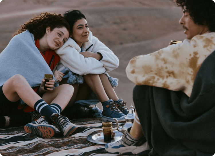 Three friends sit on patterned blanket outdoors, enjoying tea; desert landscape in background.