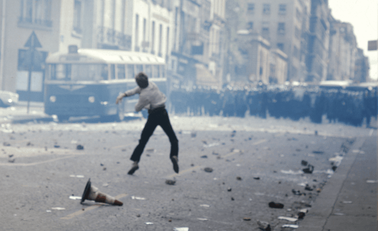 Person throws object on debris-strewn street toward riot police; buildings and bus in hazy background.