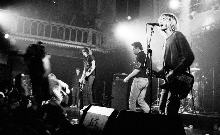 Black and white photo of band on stage singer-guitarist at mic, bright lights, crowd watching.