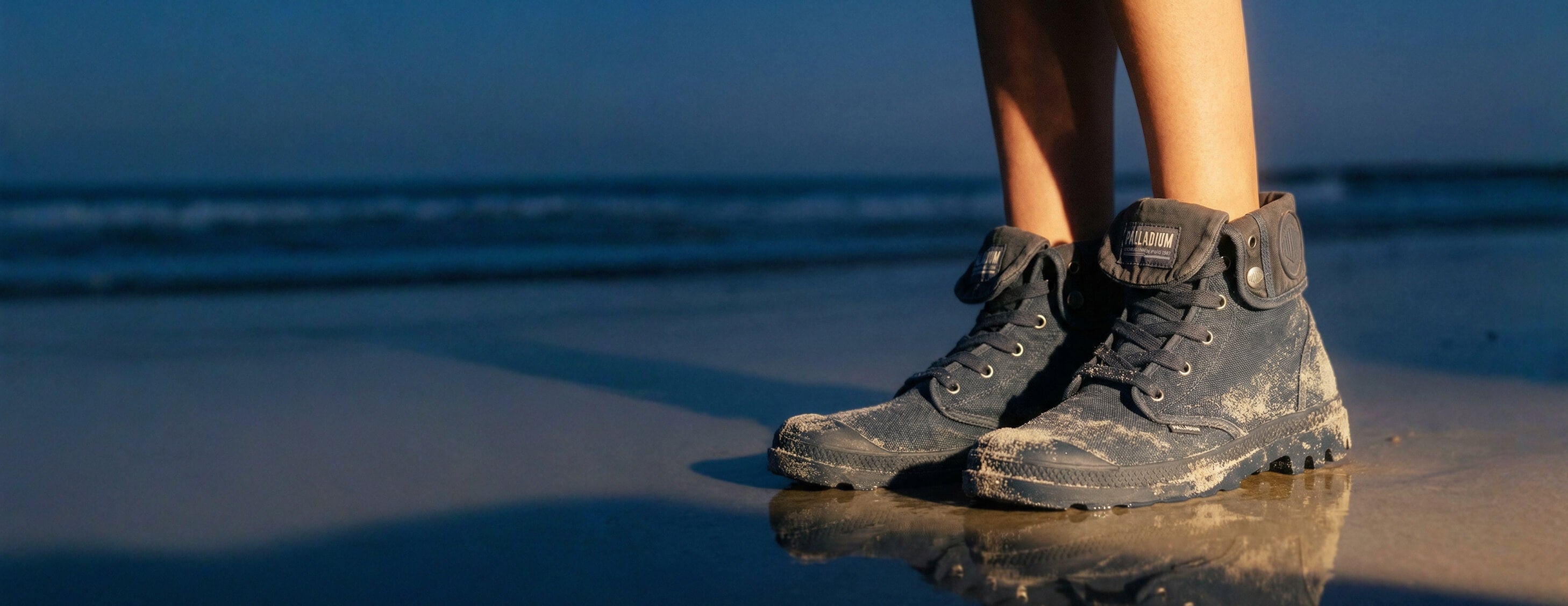 Black boots on a reflective beach surface with a blurred ocean background