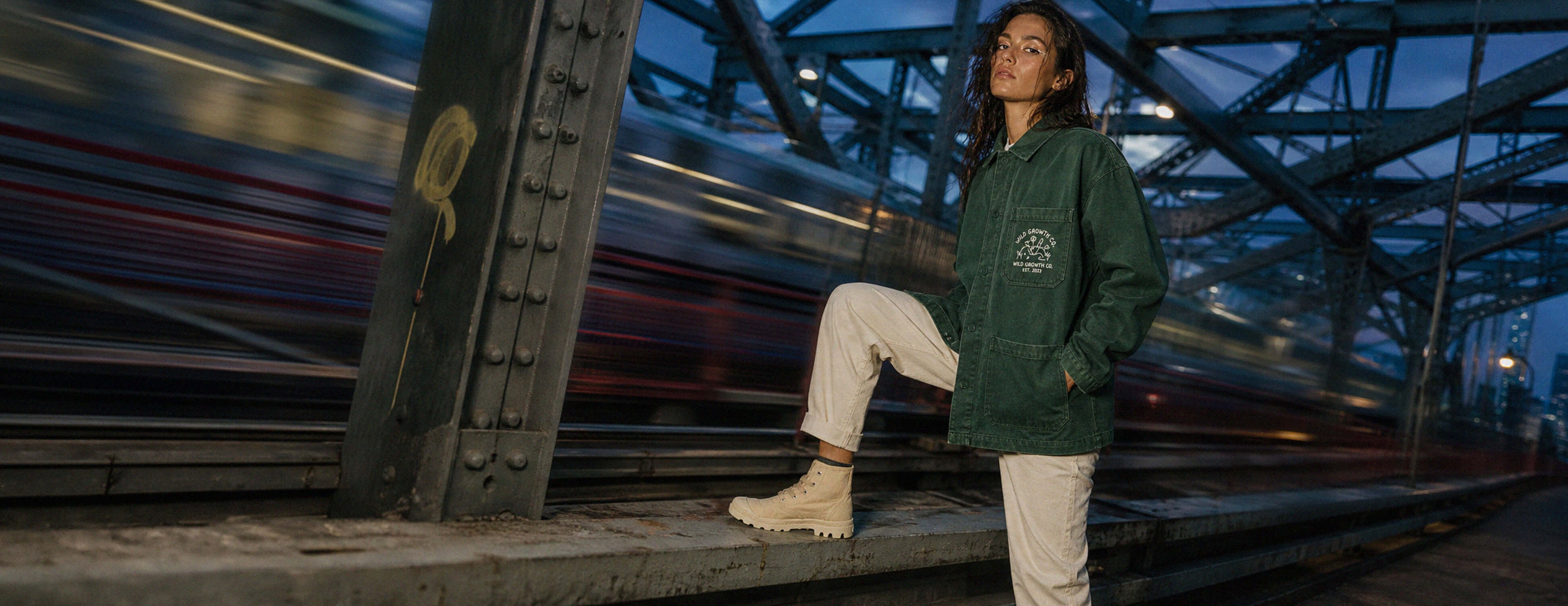 A woman stands on a bridge at dusk wearing a dark green hooded jacket, off-white pants, and off-white boots. The background shows a blurred train and steel bridge structure.