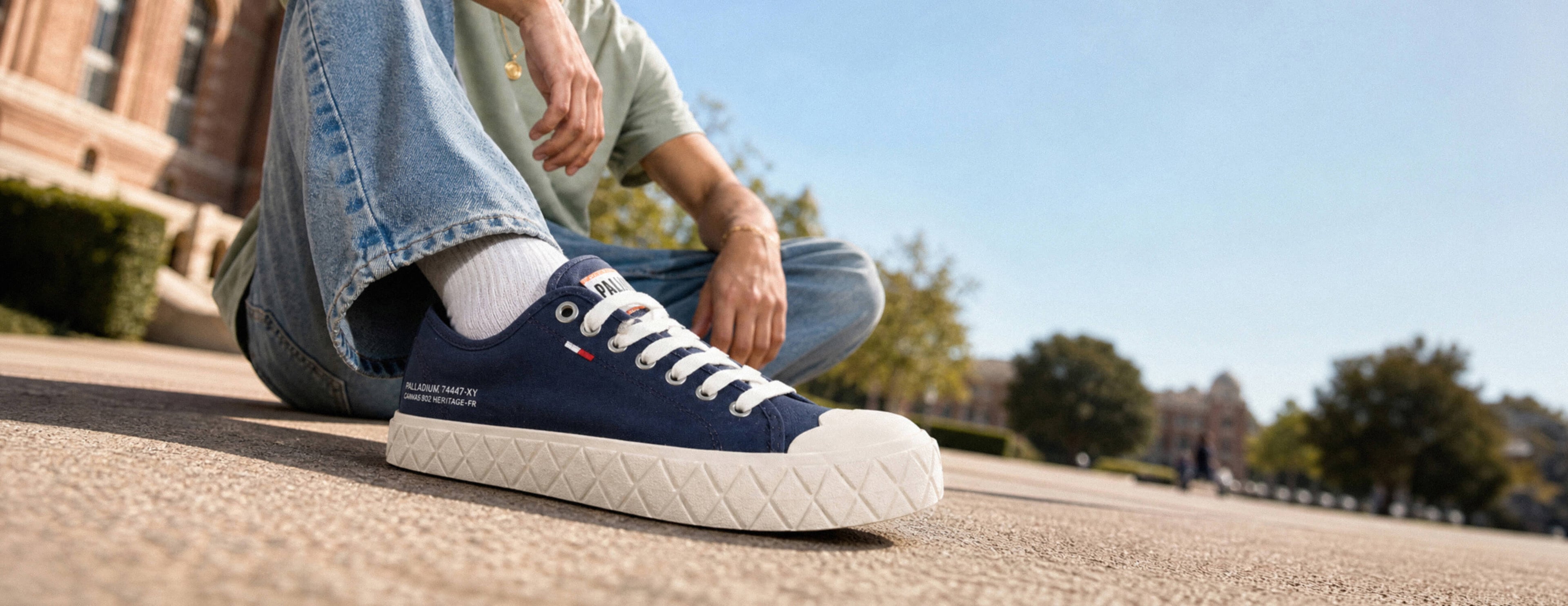 A navy blue canvas low-top sneaker with white laces and a white quilted sole, worn by a person sitting outdoors on a sunny day.