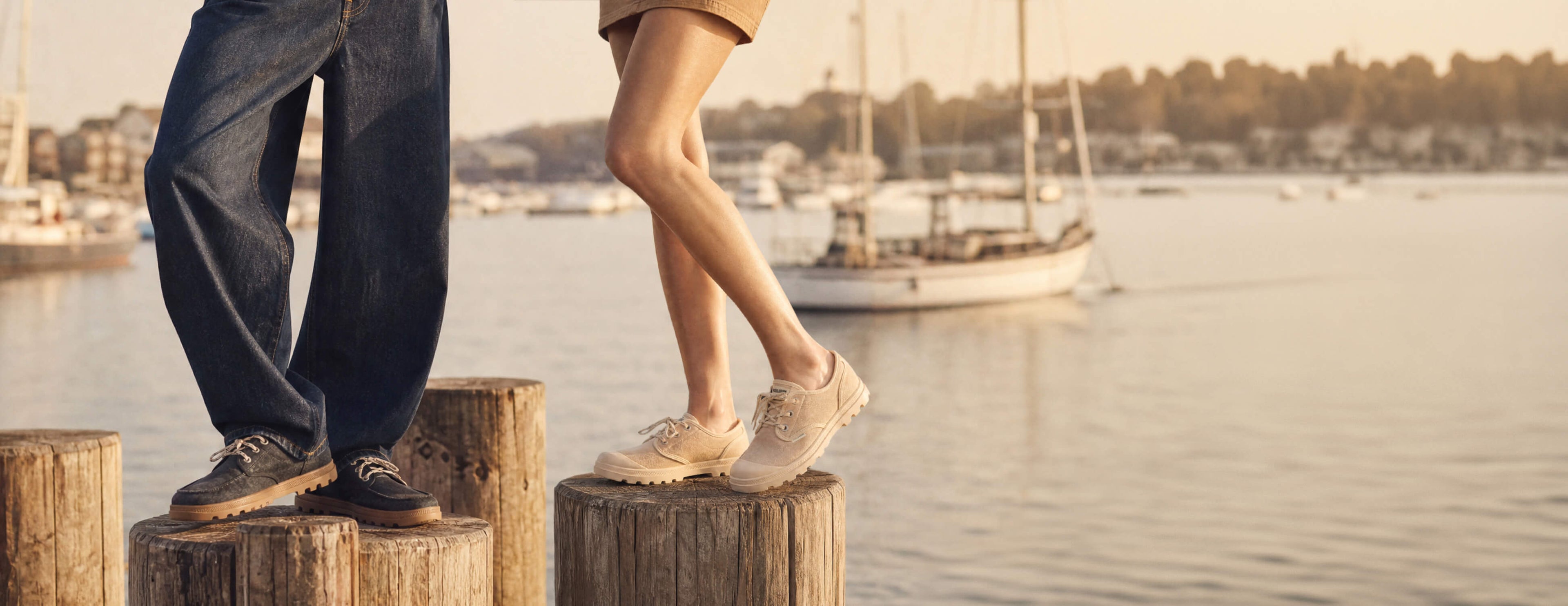 Two people standing on wooden posts by a waterfront with boats and a sunset.