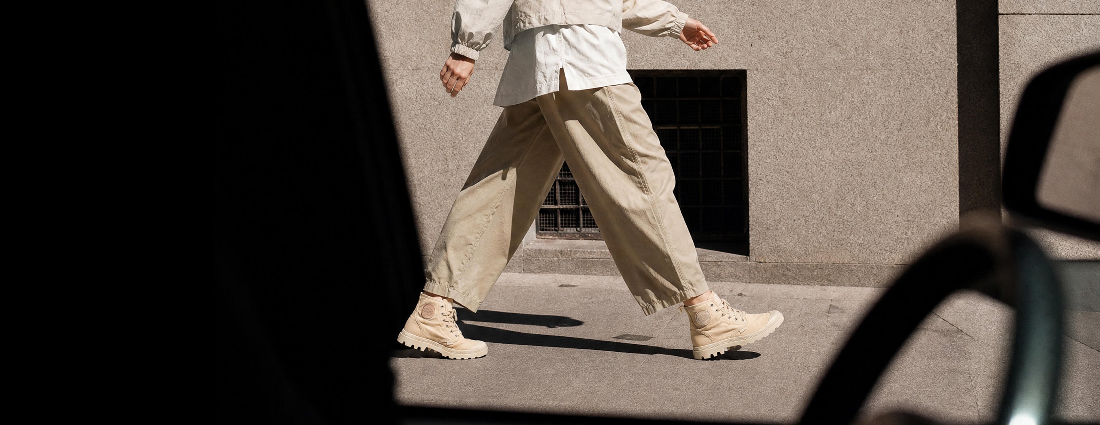 A person walking outdoors in loose beige pants, a light-colored top, and chunky beige boots, viewed through a dark foreground frame.