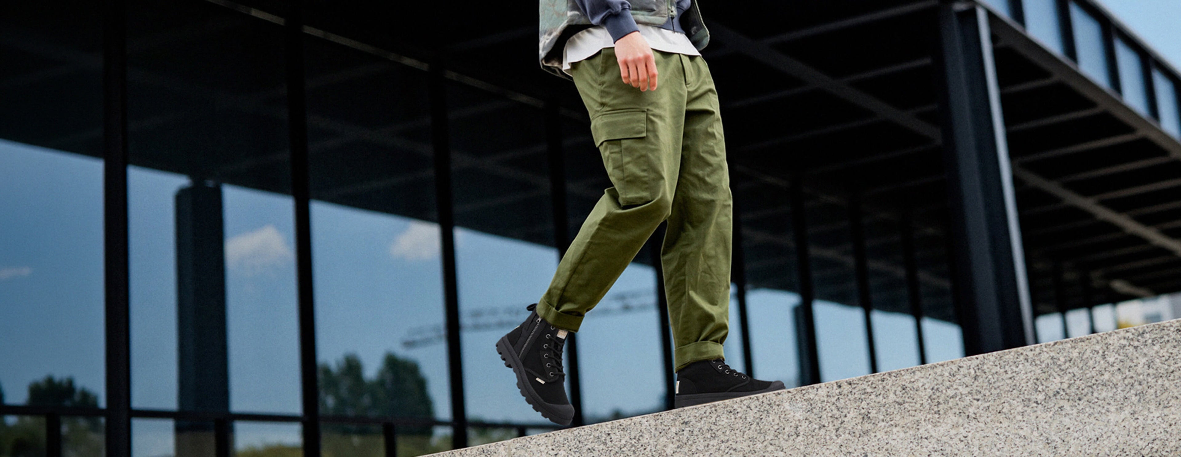 A person walking outdoors wearing olive green cargo pants, a gray jacket, and black boots in front of a modern glass building.