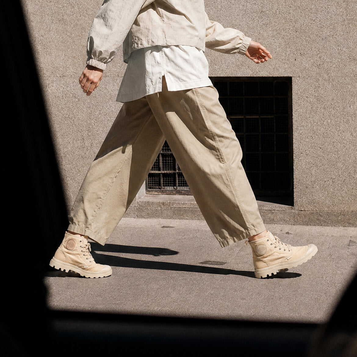 A person walking outdoors in a light beige cropped jacket, white shirt, loose beige pants, and beige high-top sneakers against a concrete wall.