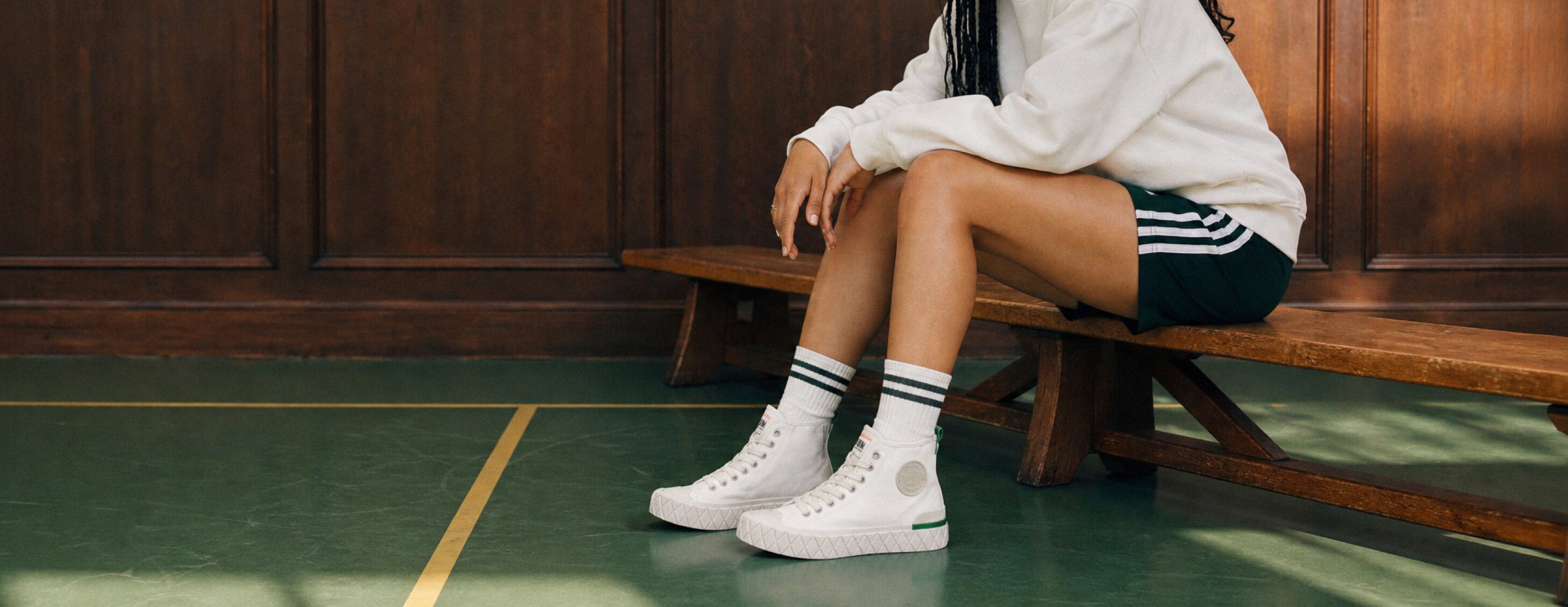 A person sitting on a wooden bench in an indoor sports hall, wearing white high-top sneakers, white crew socks with black stripes, a white sweatshirt, and dark shorts with white stripes.