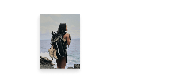 Person with long hair and backpack stands on rocky shore, gazing at sea under a cloudy sky.