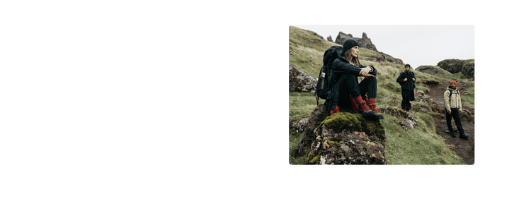 Three hikers on rocky hillside; one sits on mossy rock, two walk in the background under overcast sky.