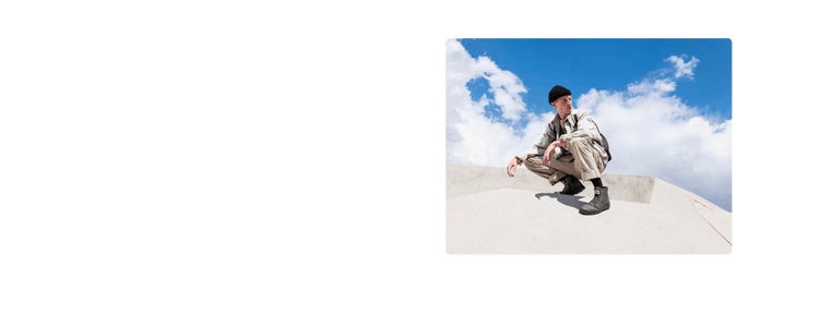 Person in beige outfit, black beanie, and boots squats on concrete structure under blue sky with clouds.
