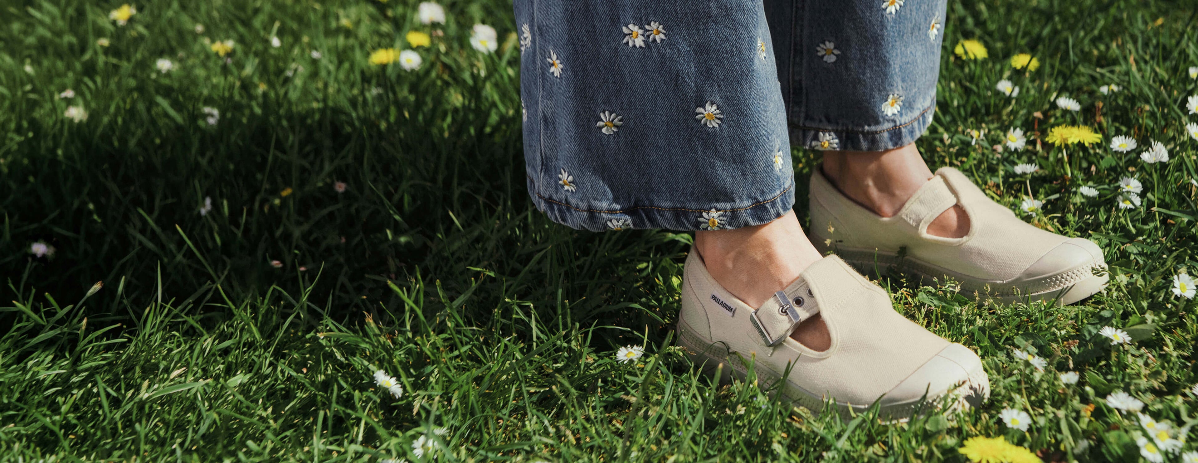 Person wearing white clogs and blue jeans with floral embroidery standing on grass with dandelions.