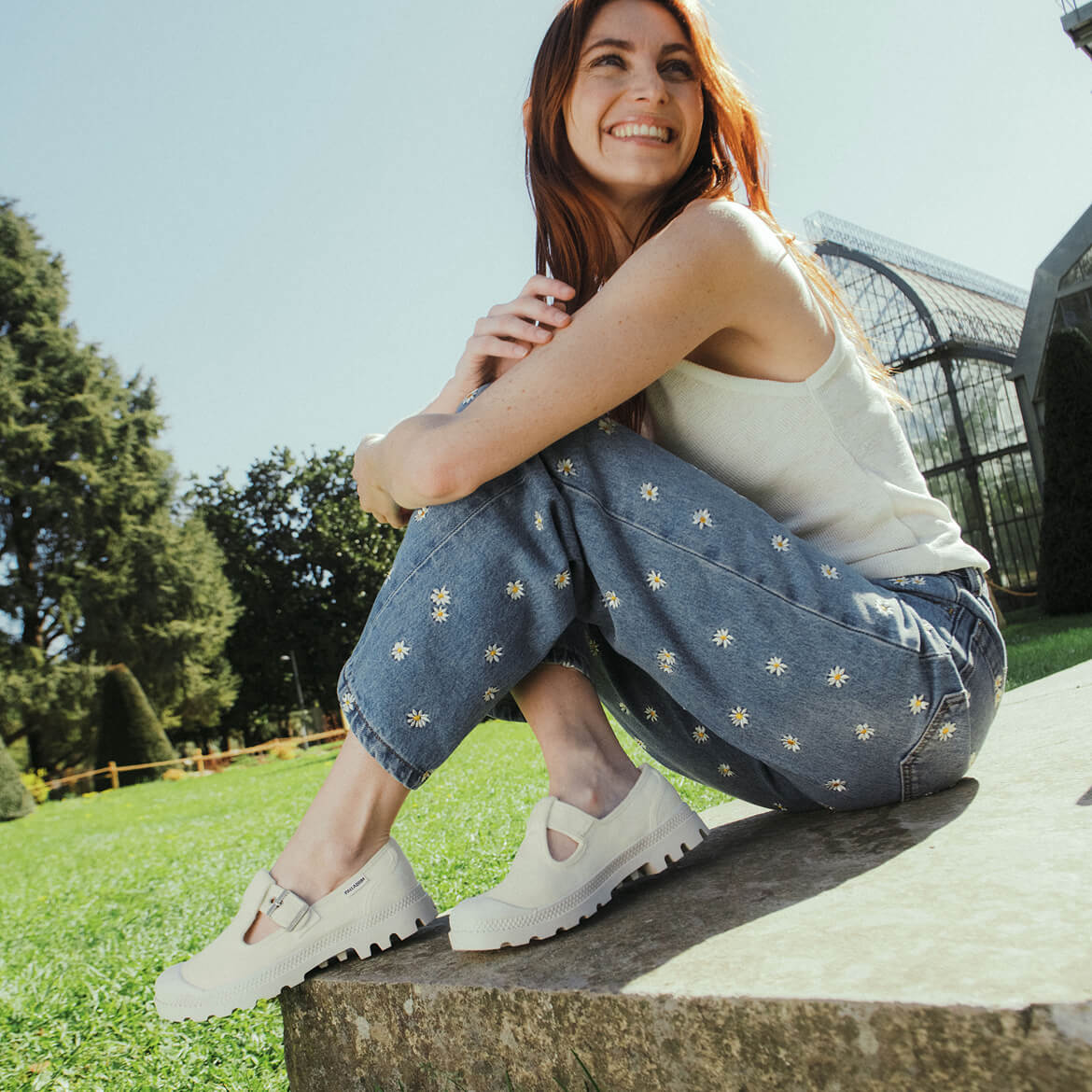 Woman sitting on a stone bench in a park wearing a white top and blue jeans with floral patterns.